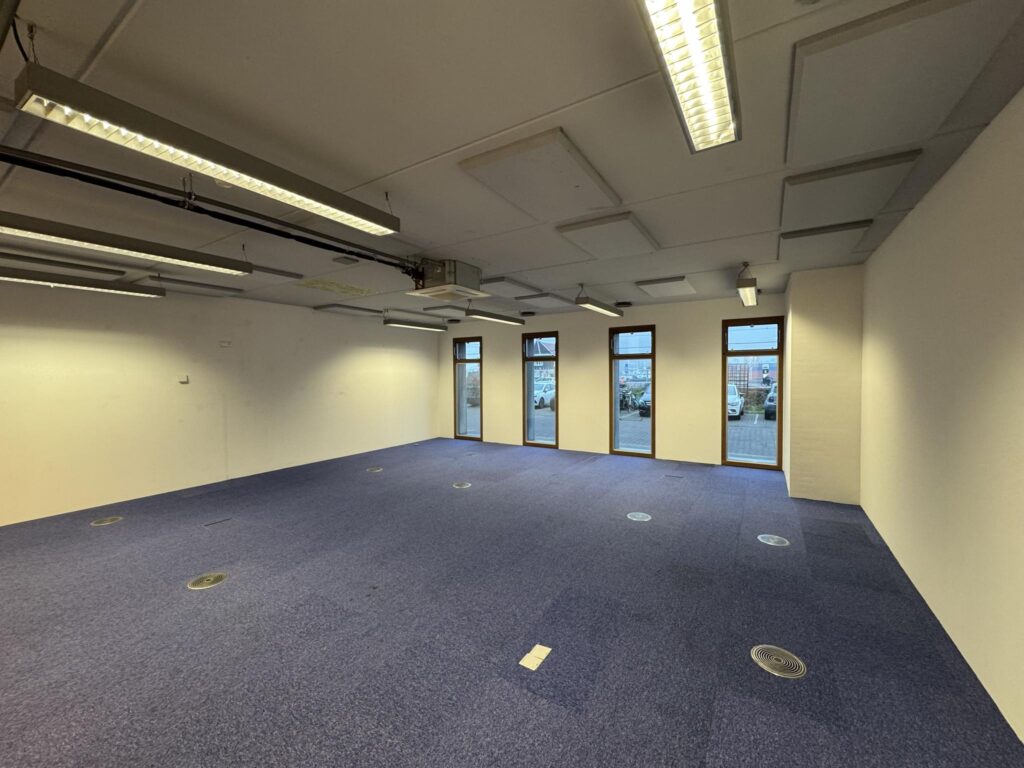 Empty office space with blue carpet, white walls, ceiling lighting, and windows facing De Ruijterkade.